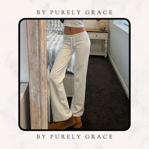 Może przedstawiać: Białe spodnie o szerokich nogawkach ze sznurkiem w pasie. Spodnie wykonane są z miękkiej, wygodnej tkaniny i mają luźny krój. Tekst "BY PURELY GRACE" jest widoczny u góry i na dole obrazu.
