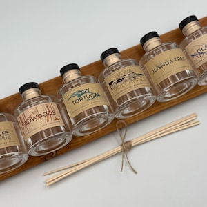 May include: Six glass bottles with cork stoppers and labels featuring different national park names and scents. The bottles are arranged on a wooden surface with a set of wooden diffuser sticks.
