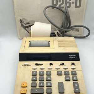 May include: Vintage Canon P26-D calculator with a cream-colored body and black accents. Features a paper feed, number keys, and function buttons. Includes a gray cord and a manual.