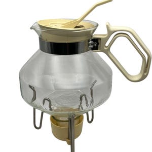 Könnte beinhalten: Vintage Kaffeekanne aus klarem Glas mit cremefarbenem Deckel, Griff und Löffel. Die Kanne hat ein silbernes Band und steht auf einem Metallständer mit einem cremefarbenen Wärmer. Ein klassisches Design.