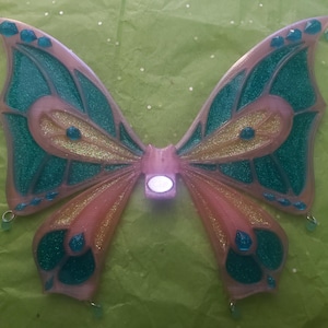 Puede incluir: Un par de alas de mariposa rosa y azul verdoso con detalles de purpurina. Las alas están unidas a una diadema y tienen una luz en el centro.