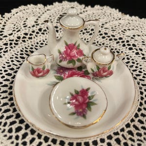 May include: A miniature porcelain tea set with a teapot, creamer, sugar bowl, and saucer. The set is white with pink rose floral designs and gold trim. The set is displayed on a white plate with gold trim.