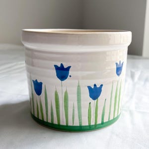 May include: White ceramic planter with a green rim and blue flower designs. The flowers are stylized tulips with green stems and leaves.