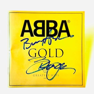 May include: A square music album cover with a bright yellow background featuring the black text ABBA, GOLD, and GREATEST HITS. Two distinct blue ink signatures are written across the centre of the cover over the printed text.