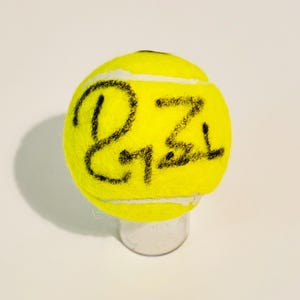 Puede incluir: Una pelota de tenis amarilla brillante con texto escrito a mano en negro. La pelota de tenis descansa sobre un pequeño soporte cilíndrico transparente. Las costuras de la pelota son blancas.