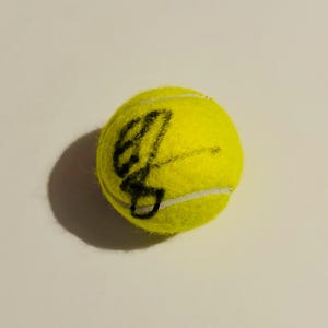 May include: A bright yellow tennis ball with a black handwritten design. The ball has white seams and a fuzzy texture. The tennis ball is on a white surface, casting a shadow.