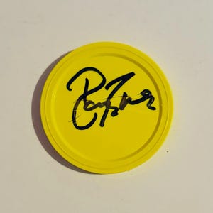 May include: A bright yellow, circular lid with a black handwritten signature. The lid has a raised edge and a smooth surface. The signature appears to be a stylized name or phrase.