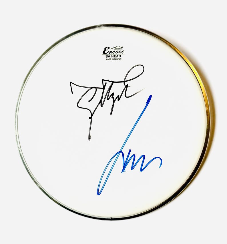 Puede incluir: Un parche de tambor blanco circular con un borde met&aacute;lico, que presenta el texto negro Encore by Remo BA HEAD MADE IN TAIWAN en la parte superior. La superficie muestra dos firmas distintas: una en tinta negra y otra en tinta azul en el centro.