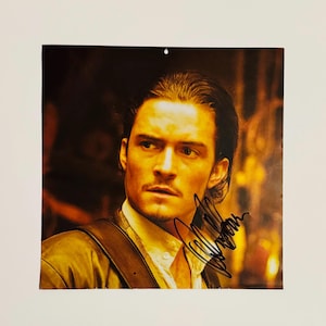 Puede incluir: Una fotografía en blanco y negro de un hombre con cabello oscuro y barba. Lleva una chaqueta de cuero marrón y una camisa blanca. La fotografía está firmada con el nombre "Orlando Bloom".