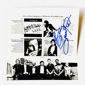 Puede incluir: Un inserto de papel blanco con un collage en blanco y negro de portadas de álbumes de Bruce Springsteen y una foto de la banda, con un autógrafo en tinta azul firmado en el lado derecho. El texto enumera títulos, años y listas de canciones.