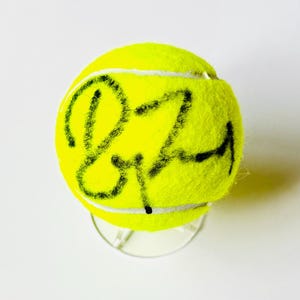 Puede incluir: Una pelota de tenis de color amarillo neón brillante con un autógrafo en tinta negra escrito sobre la superficie difusa. La pelota descansa sobre un soporte de exhibición circular de plástico transparente, resaltando la firma en la textura.