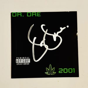 Puede incluir: Portada de álbum negra con el texto "DR. DRE" en verde en la parte superior. Una firma blanca está en el centro, con una hoja de cannabis verde y el número "2001" en la parte inferior. La portada también tiene una advertencia de "Parental Advisory Explicit Content".