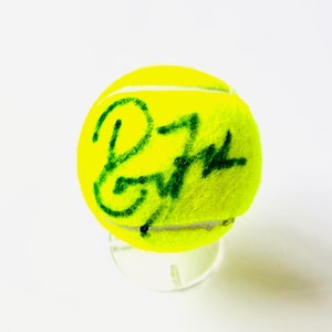 Puede incluir: Una pelota de tenis de color amarillo neón brillante con una textura de fieltro difusa y una costura de goma blanca. La superficie de la pelota está marcada con una firma o autógrafo escrito a mano en verde oscuro que cruza el centro.