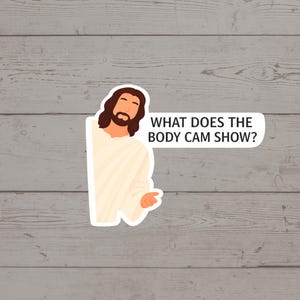 May include: A sticker featuring a cartoon illustration of Jesus peeking from behind a white shape. The text "WHAT DOES THE BODY CAM SHOW?" is in a white speech bubble. The background is a gray wooden surface.