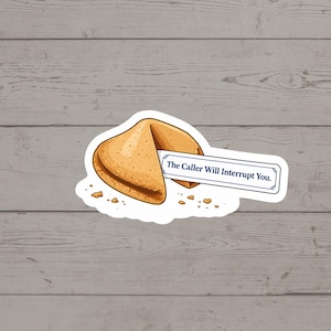 May include: A sticker featuring a cartoon fortune cookie with a message. The cookie is light brown with darker brown edges and a white border. The fortune reads, "The Caller Will Interrupt You." Small crumbs are scattered around the cookie.