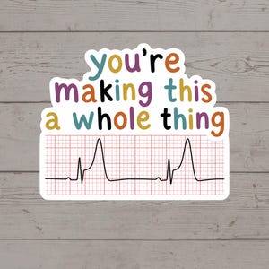 May include: A sticker with the text "you're making this a whole thing" in various colors. Below the text is a black line representing an EKG, on a red grid background. The sticker is on a light wood grain surface.