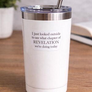 Op de afbeelding: Witte beker met een roestvrijstalen rand en een doorzichtig deksel. De beker heeft een roestvrijstalen rietje en een sticker met de tekst: "I just looked outside to see what chapter of REVELATION we're doing today."