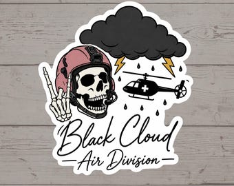 Puede incluir: Una pegatina con una calavera que lleva un casco de vuelo rosa y hace el signo de la paz. Sobre la calavera hay una nube de lluvia oscura con rayos amarillos y un helicóptero negro con una cruz blanca. El texto dice Black Cloud Air Division.