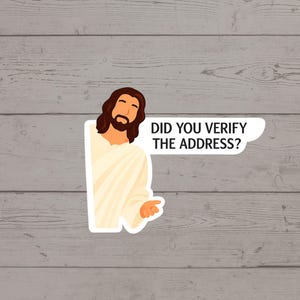 May include: A sticker featuring a cartoon illustration of Jesus in a white robe, with brown hair and a beard. A speech bubble says, "DID YOU VERIFY THE ADDRESS?" The sticker is on a gray wooden background.
