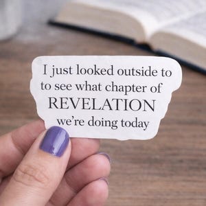 Op de afbeelding: Witte sticker met de tekst "I just looked outside to see what chapter of REVELATION we're doing today." De sticker wordt vastgehouden door een hand met paarse nagellak. Op de achtergrond is een boek te zien.