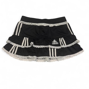 May include: Black Adidas skirt with white stripes and a white lace trim. The skirt has a tiered design with the Adidas logo on the front. The waistband is black with a silver stripe.