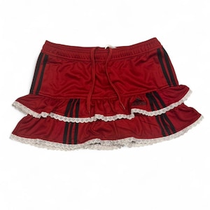 May include: A red Adidas skirt with black stripes down the sides. The skirt has a drawstring waist and two ruffled layers with white lace trim. The Adidas logo is visible on the skirt.