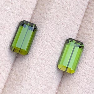 以下が含まれることがあります： 鮮やかなオリーブグリーンの色合いをした、エメラルドカットの長方形の宝石2つ。それぞれの石には光を反射する正確な直線状のファセットが施されており、結晶構造全体に明確な縦線が見える、透明感のある外観を作り出しています。