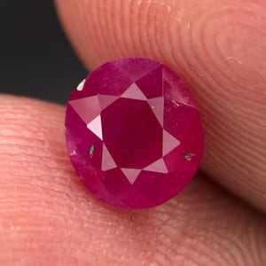 Puede incluir: Una piedra preciosa redonda y facetada de color rojo intenso y vibrante. La piedra presenta un patrón simétrico en forma de estrella de facetas triangulares que irradian desde el centro, con pequeñas inclusiones naturales oscuras visibles.