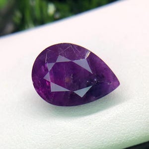 Puede incluir: Una piedra preciosa de color púrpura oscuro en forma de pera con un corte facetado. La piedra preciosa tiene un patrón en forma de estrella en el centro.