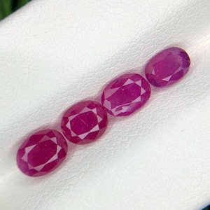 May include: Four oval-shaped gemstones, each a vibrant pink hue, are arranged in a row on a white surface. The gemstones are smooth and polished, with a slight sparkle.