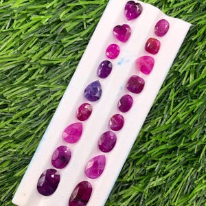 May include: A collection of faceted loose gemstones in various shades of pink, purple, and magenta, arranged in two vertical rows on a white display card. The stones feature diverse shapes including ovals, pears, hearts, and cushion cuts.