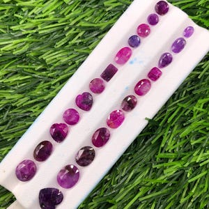 May include: A collection of loose faceted gemstones in various shades of purple, pink, and magenta, arranged in two parallel rows on a white display pad. The stones feature diverse shapes including ovals, rounds, hearts, and a single rectangular cut.