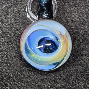 May include: A round, glass pendant with a swirling blue and yellow design, resembling a galaxy. The pendant has a black, glittery top and a clear loop for a chain. The design includes a white line and flecks of glitter.
