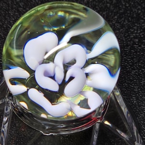 Può includere: Una biglia di vetro trasparente con forme astratte bianche e vorticose. Il marmo ha una tonalità verde e blu ed è esposto su un supporto di vetro trasparente. Il design assomiglia a un fiore.