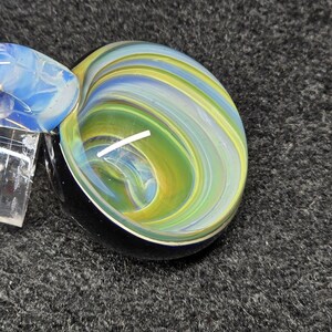 Op de afbeelding: Een handgeblazen glaskunstwerk met een wervelend patroon van groen, geel, blauw en wit. Het stuk heeft een zwarte basis en een heldere, rechthoekige bevestiging. De kleuren vloeien samen in een spiraalvormig ontwerp.