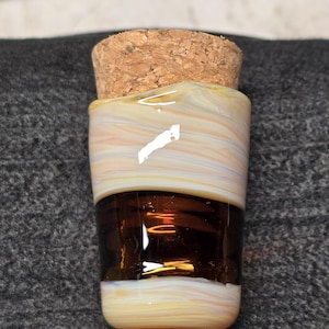 Peut inclure: Un petit récipient en verre avec un bouchon en liège, présentant une bande centrale ambre foncé contrastée par des sections supérieure et inférieure aux motifs de verre irisé blanc, pêche et jaune pâle avec une finition lisse.