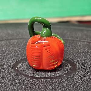 May include: A small, handcrafted glass pumpkin with a vibrant orange body and a green stem. The pumpkin has detailed vertical lines and a loop handle. The background is a blurred green.