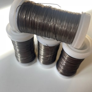 Può includere: Quattro bobine di filo artigianale marrone scuro sono impilate, con bobine di plastica bianca. Il filo è avvolto strettamente e le bobine sono disposte ad angolo, creando un display visivamente accattivante. Il filo sembra essere di colore metallico.