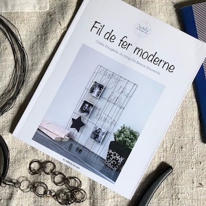 Op de afbeelding: Een boek getiteld "Fil de fer moderne" van Claire Rougerie, met een draadraster met foto's en de zin "enjoy the little things". Zwart draad en een decoratieve ketting zijn ook zichtbaar.