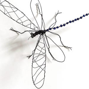 May include: A handmade wire dragonfly sculpture. The dragonfly is made of black wire with detailed wings. The tail has small, blue beads. The sculpture is set against a white background.