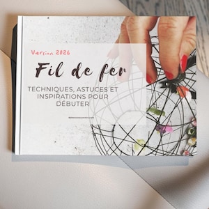Può includere: Un libro intitolato "Fil de fer" con il testo "Version 2026" in rosa. La copertina del libro presenta una scultura di filo metallico e il testo "TECHNIQUES, ASTUCES ET INSPIRATIONS POUR DÉBUTER". Il libro è su una superficie bianca.