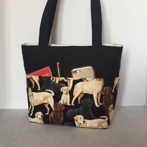 May include: Black tote bag with a zippered pocket and a phone pocket. The bag is lined with a cream fabric and has a pattern of Labrador Retrievers in black, brown, and yellow on the front.