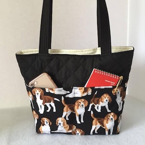 May include: Black quilted tote bag with a beige lining and a front pocket. The bag features a black and white print of beagle dogs. The pocket contains a red memo book and a gold pen.