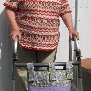 K&ouml;nnte beinhalten: Eine Frau mit wei&szlig;en Haaren benutzt einen Rollator mit einer floral gemusterten Tasche. Sie tr&auml;gt ein rot-wei&szlig;-braunes gestreiftes Shirt und eine beige Hose.