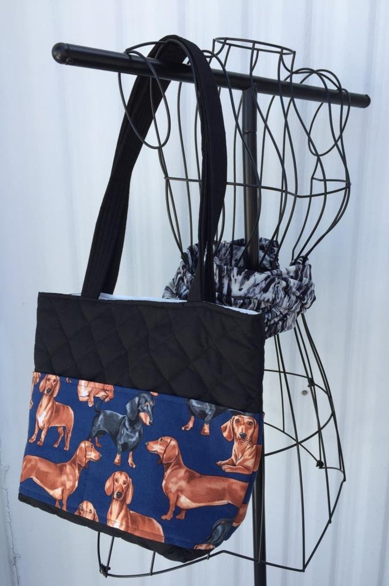 May include: Black quilted tote bag with long black straps. The bottom section features a blue fabric printed with dachshunds in various poses. A black wire mannequin and a black and white scarf are in the background.