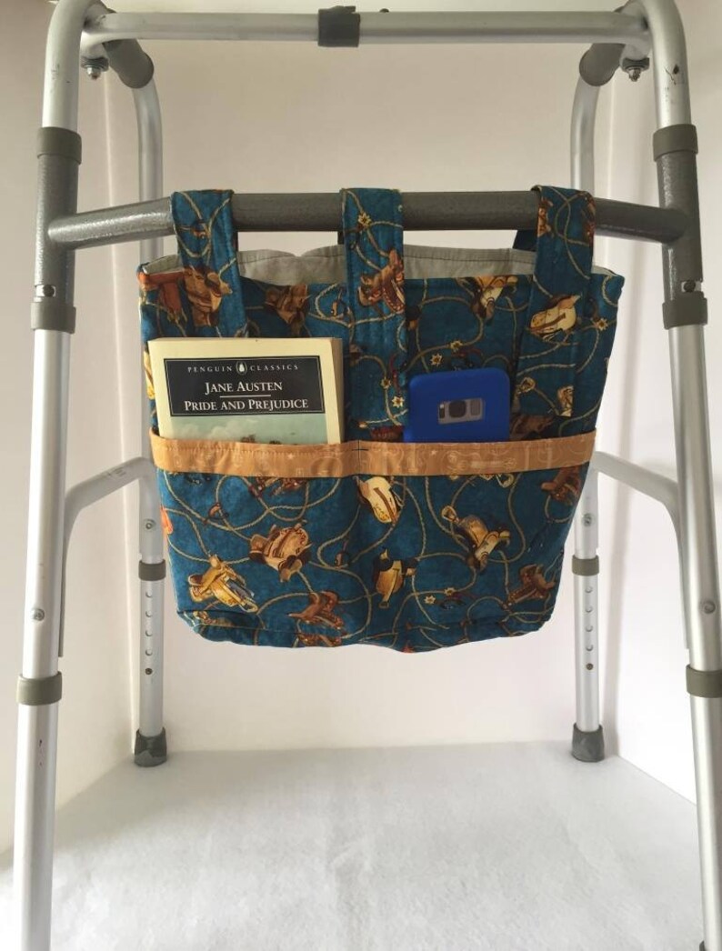 K&ouml;nnte beinhalten: Eine blaue und braune Stofftasche mit Western-Design h&auml;ngt an einem Rollator. Die Tasche hat mehrere F&auml;cher und enth&auml;lt ein Buch mit dem Titel "Stolz und Vorurteil" von Jane Austen und ein blaues Telefon.