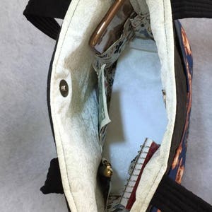 May include: Open view of a black tote bag with black handles. The interior features a floral patterned lining and several pockets. A cell phone, cash, and a small notebook are visible inside the bag.
