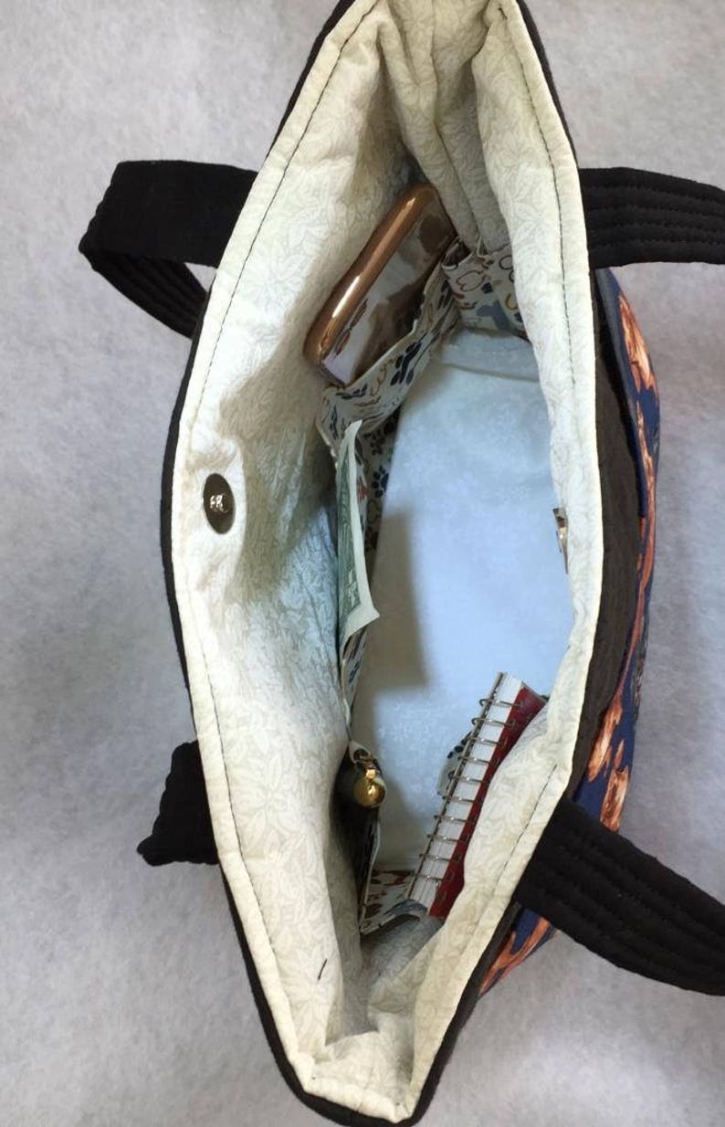 May include: Open view of a black tote bag with black handles. The interior features a floral patterned lining and several pockets. A cell phone, cash, and a small notebook are visible inside the bag.
