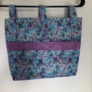 May include: A blue and purple tote bag with a floral print and a purple suede trim. The bag has two straps and is hanging on a black hanger.