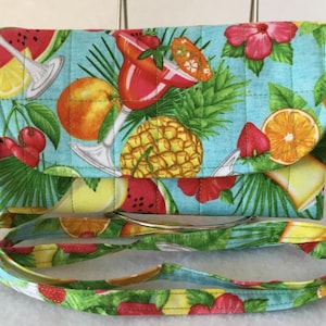 Pode incluir: Uma bolsa de ombro acolchoada turquesa com estampa de frutas tropicais. A estampa inclui abacaxi, melancia, laranjas, cerejas e uma ta&ccedil;a de margarita. A bolsa tem uma al&ccedil;a verde e amarela.